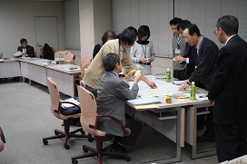 8人の参加者が1つのテーブルの周りに集まり、手元の付箋を見ている方やテーブルの上に置かれた模造紙を指さしている方がいるグループ写真