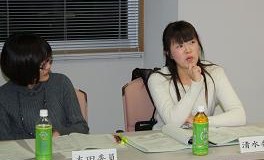 参加者の女性2人が席に着き、右側の白い服を着た女性が右手をあごの下に添えて話している様子を左側に座っている女性が見ている写真